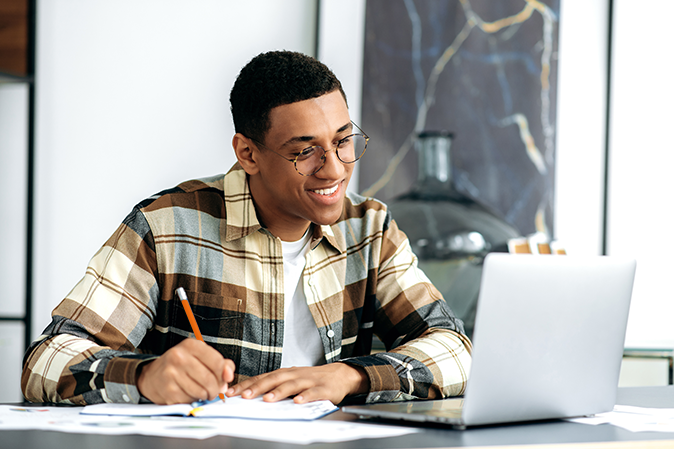 Online education, distant learning. Hispanic attractive positive guy with glasses, uses a laptop, takes notes during an online lesson, sits at a desk, stylishly dressed, smiling, gains knowledge