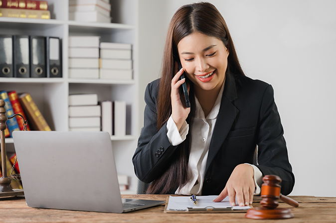 Confident Lawyer on a Call: A portrait of a smiling Asian female lawyer in her office, confidently reviewing documents while on a phone call, showcasing professionalism and success.