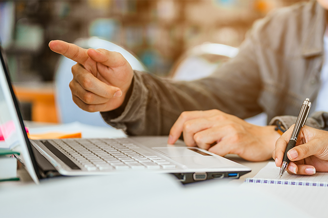 Man pointing at laptop screen while a woman takes notes