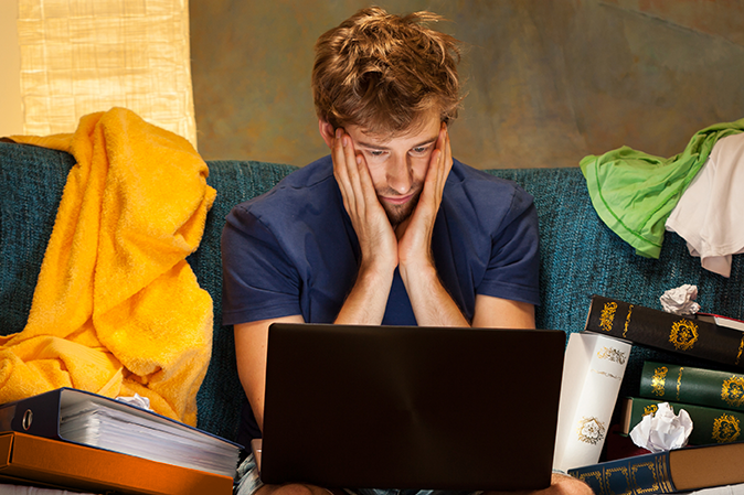 Frustrated young man holding his head looking at his laptop in his messy room