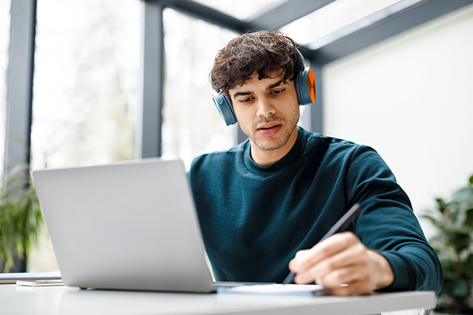 Young man wearing headphones taking notes in front of laptop