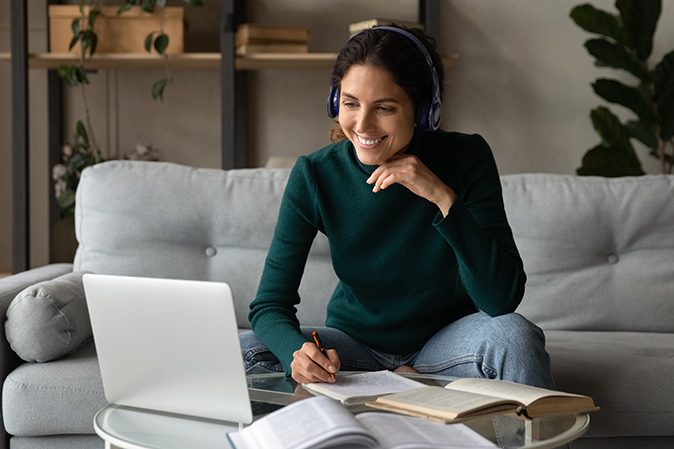 Smiling woman in headphones studying online at home, looking at laptop screen, watching webinar, involved in internet lesson, listening to lecture, motivated positive female student taking notes